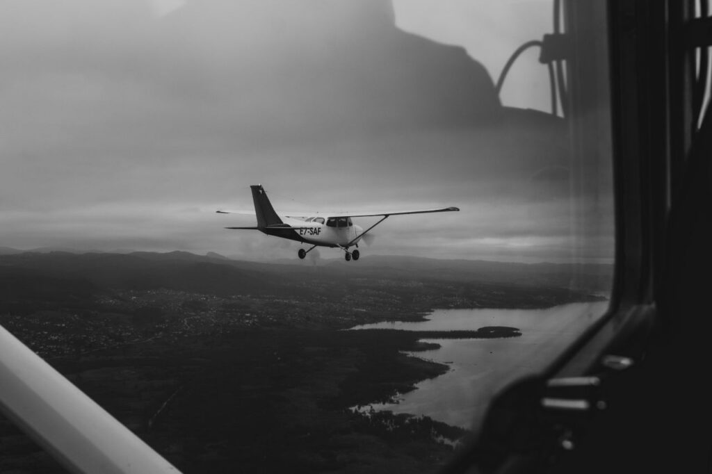Small airplane flying over a landscape beneath a cloudy sky, viewed from another aircraft cockpit.