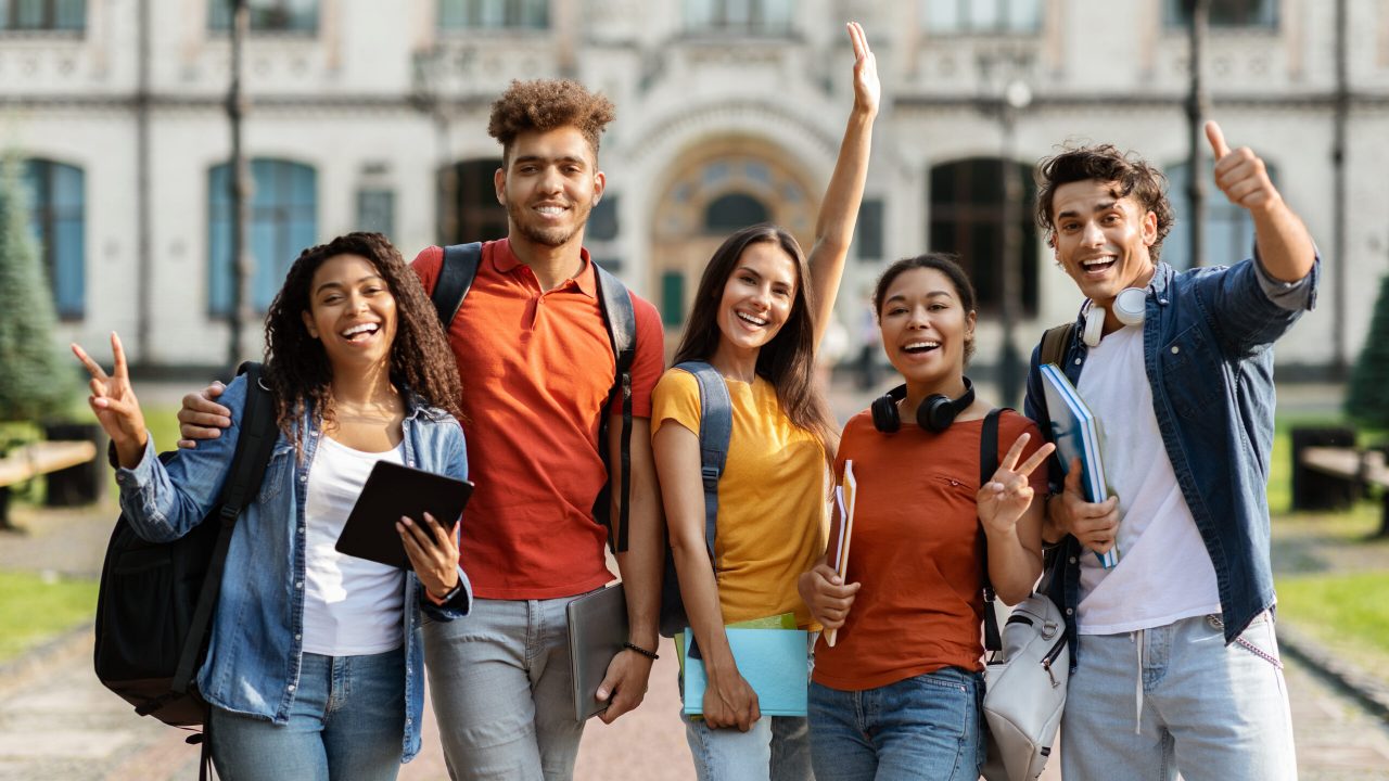 Freshmen Orientation Concept. Group Of First Year Students With Workbooks Posing Together Outdoors, Happy Multiethnic Young People Standing Near University Building And Smiling At Camera